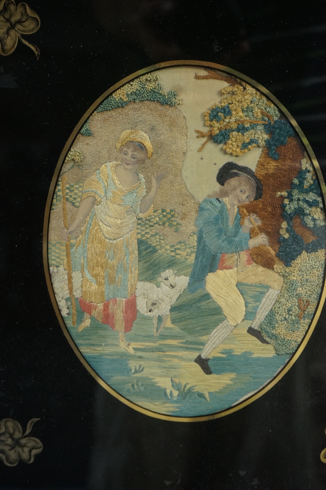 Two early 19th century oval silk worked embroideries. Both figural pastoral scenes, mostly worked in stem stitch, the leaves in a stump work style knotting. Both embroideries set within decorative Verre Eglomise glass an